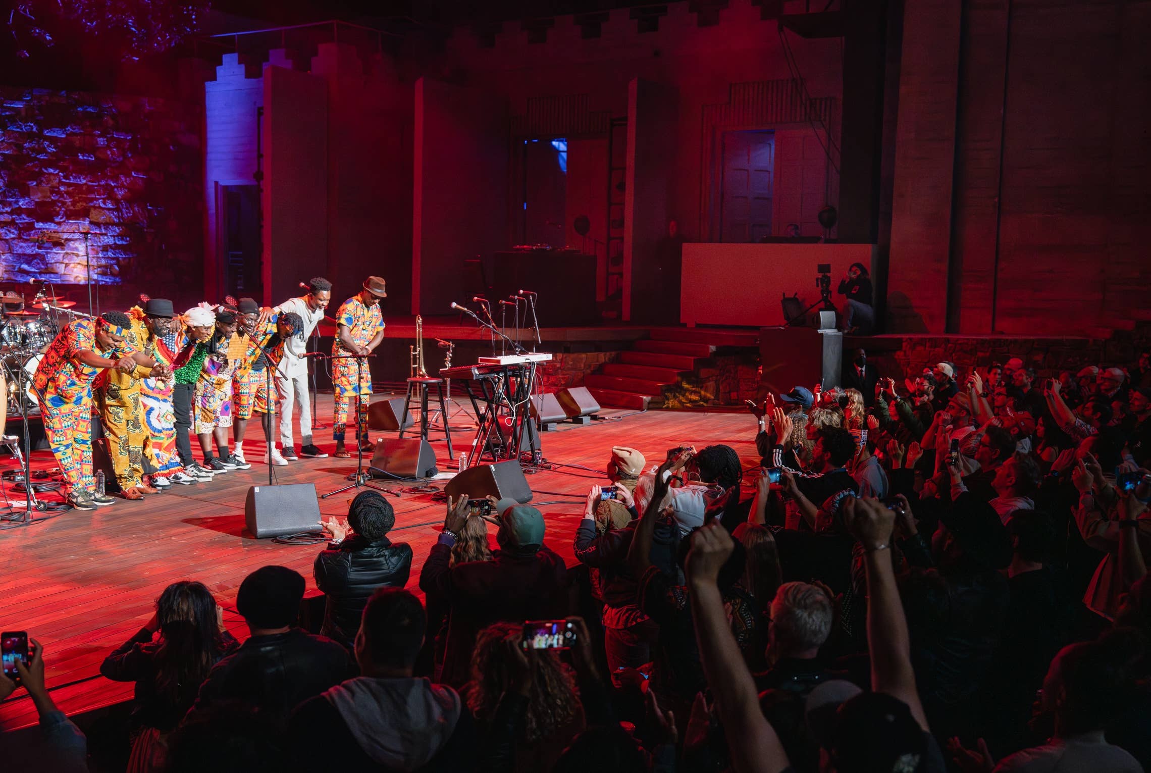 Band members in colorful outfits perform on stage as the crowd cheers under red and blue lighting
