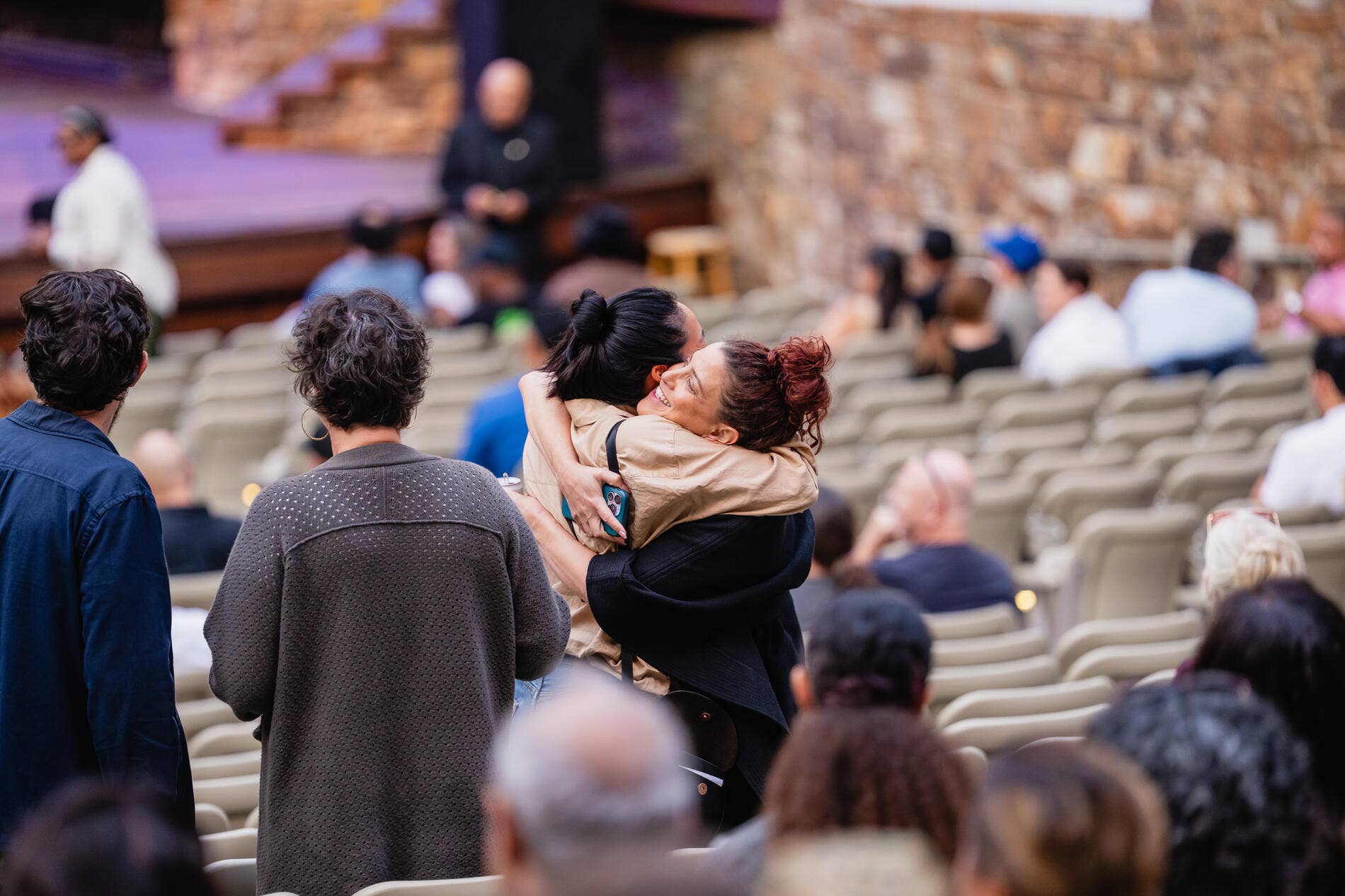Woman hugs seated man in stadium seats as eventgoers fill the background rows