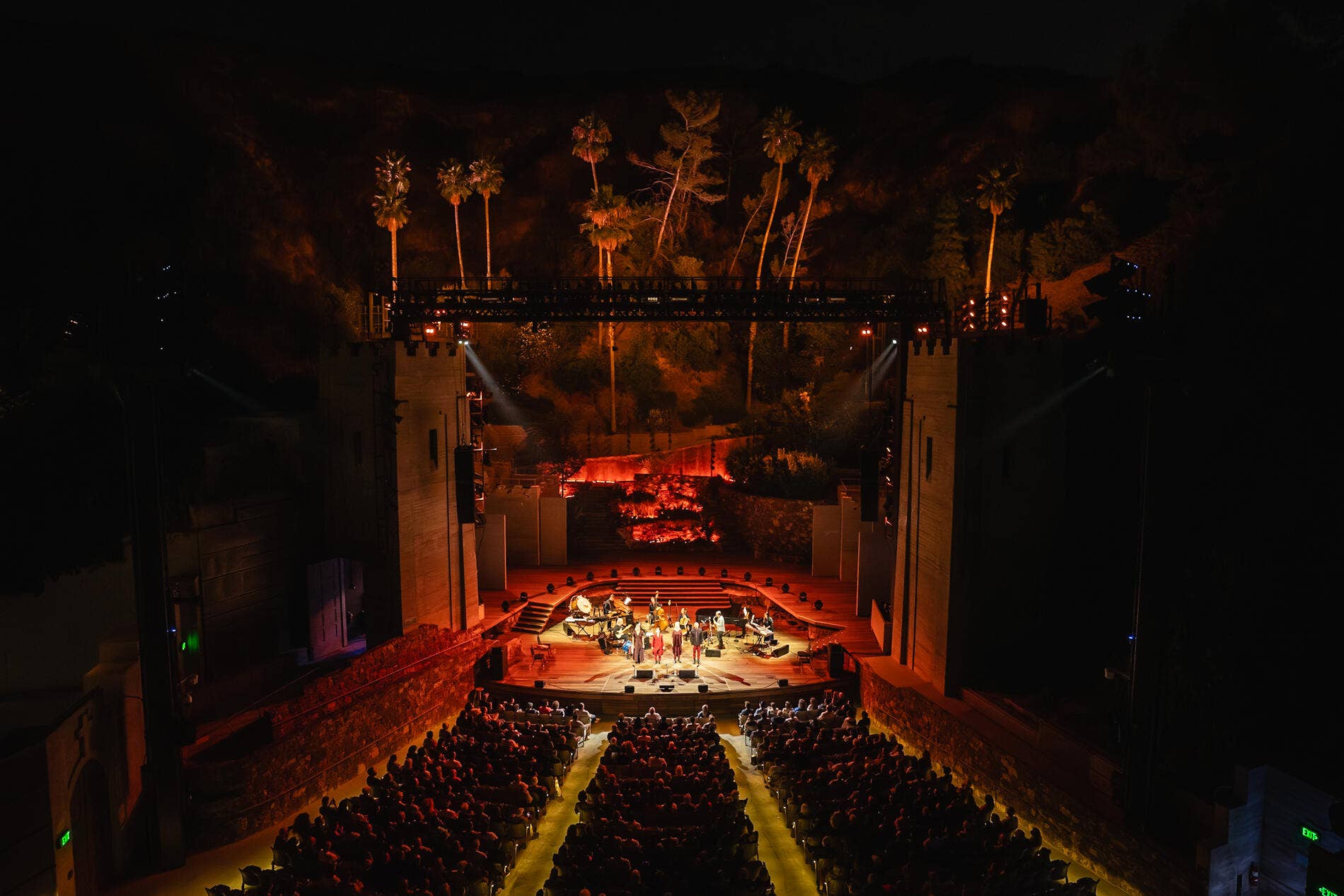 Theater stage at night with musicians and an audience, lit by warm spotlights and hanging palm trees on backdrop.
