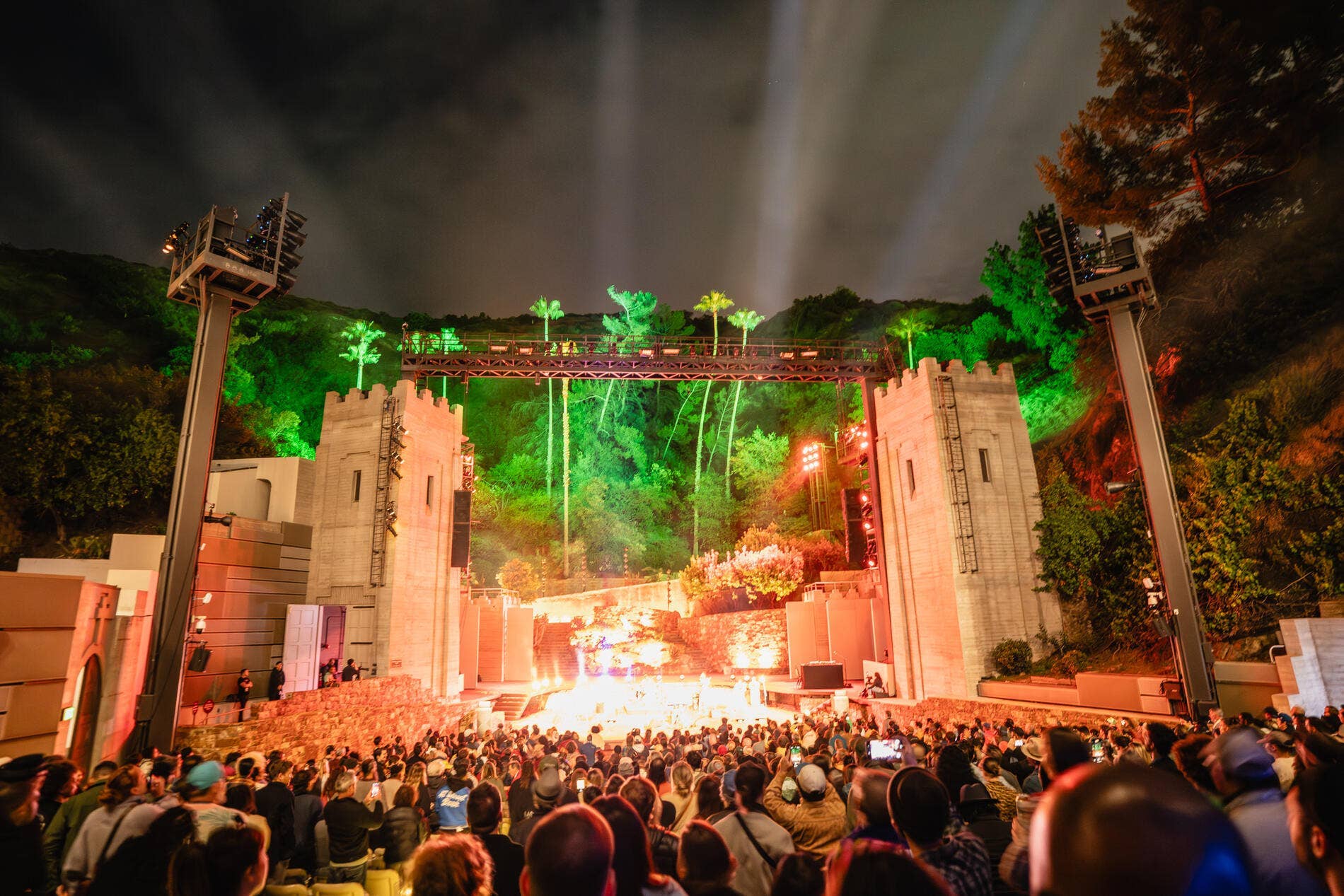Outdoor concert stage at night with green and red spotlights, palm décor, and a large cheering crowd in front