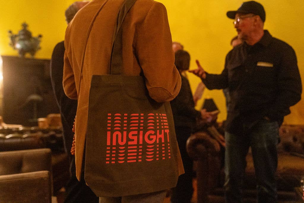 Man in a tan hoodie and tote bag with red “InSight” logo, walking outdoors as people speak nearby.