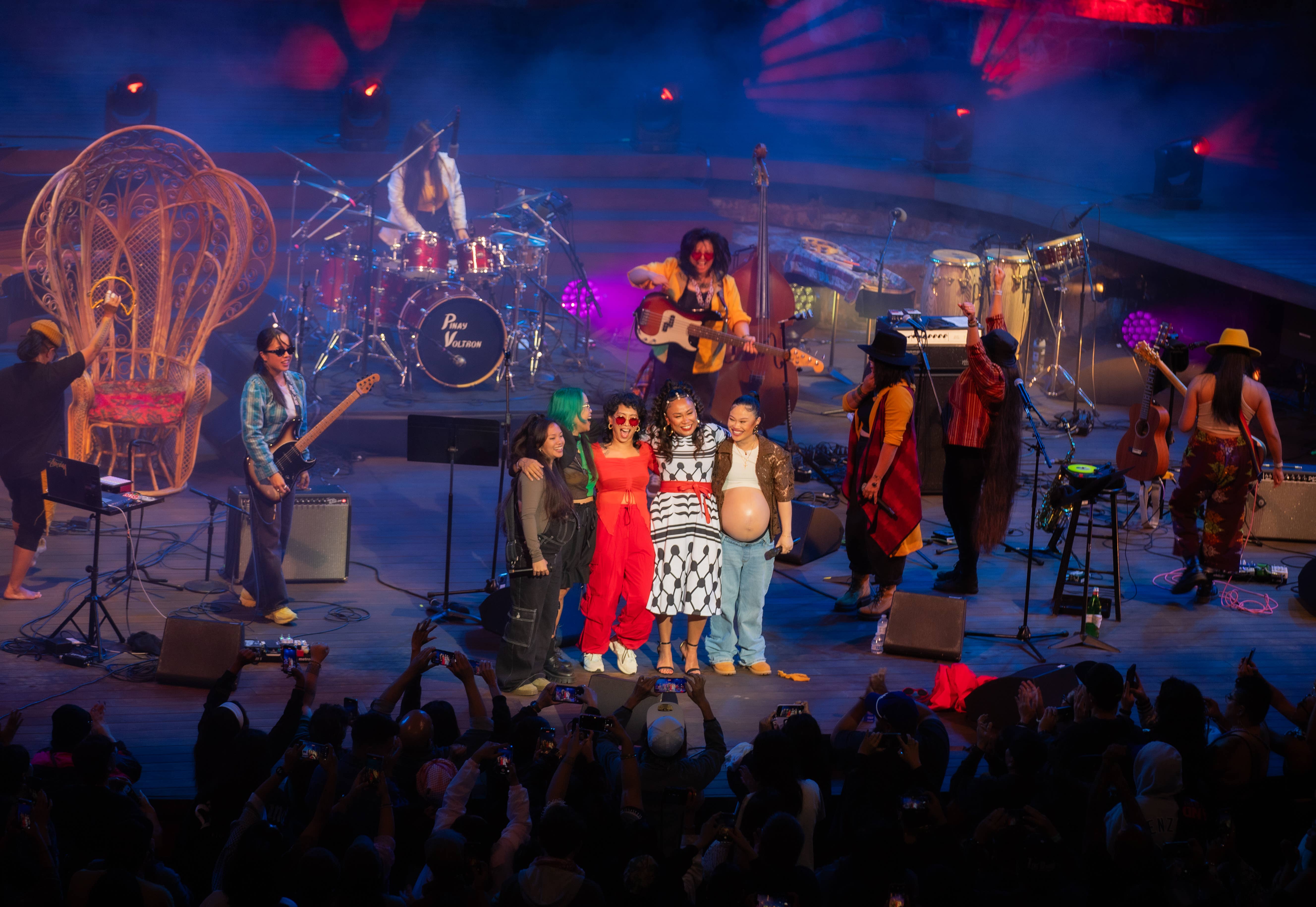 Band performing on stage with colorful lighting, brass instruments, and a cheering crowd holding phones.