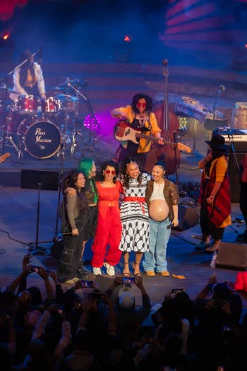Band performing on stage with colorful lighting, brass instruments, and a cheering crowd holding phones.