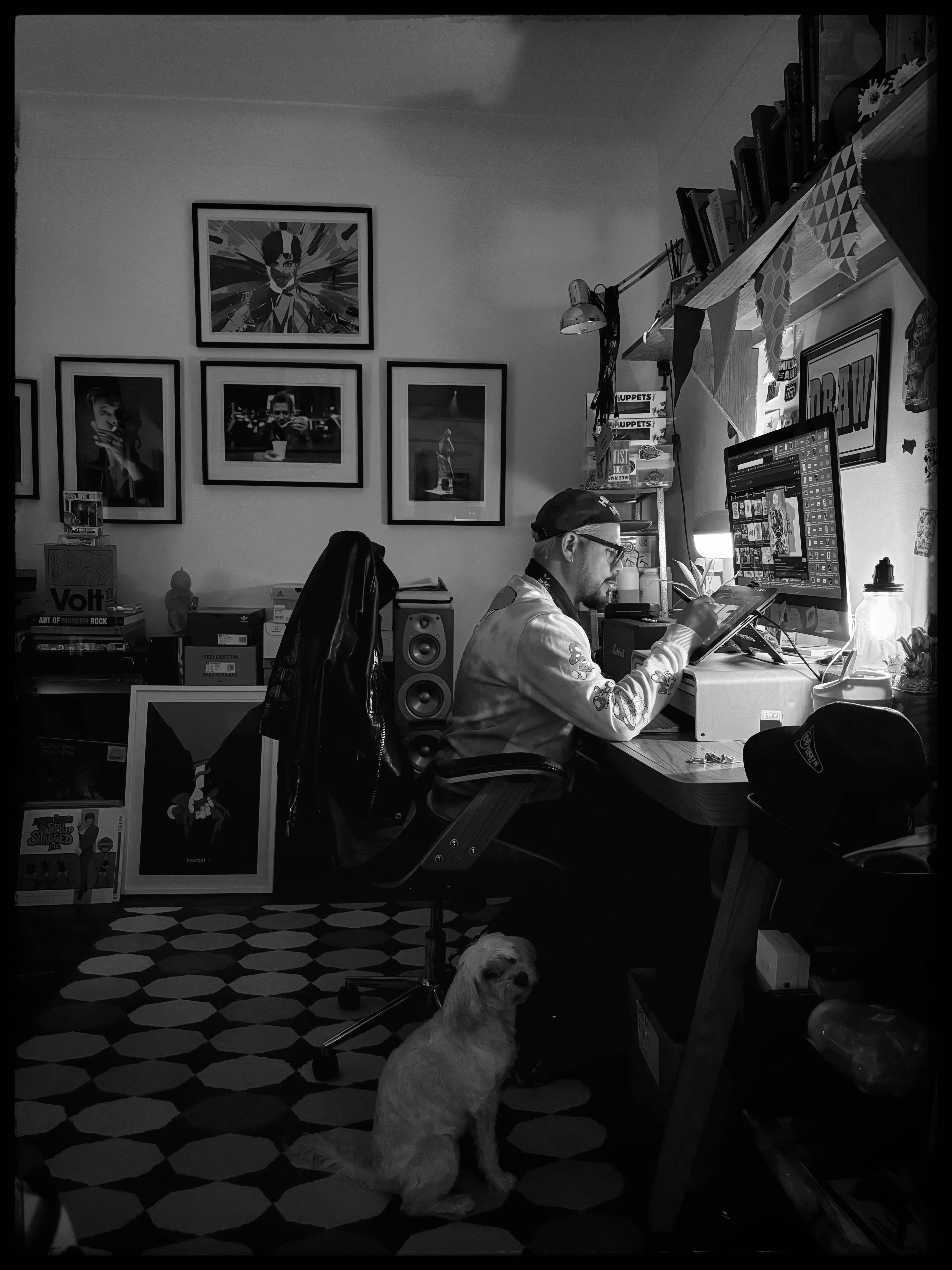 Man seated at computer editing, dog on floor, framed art and books on wall in a black-and-white studio.