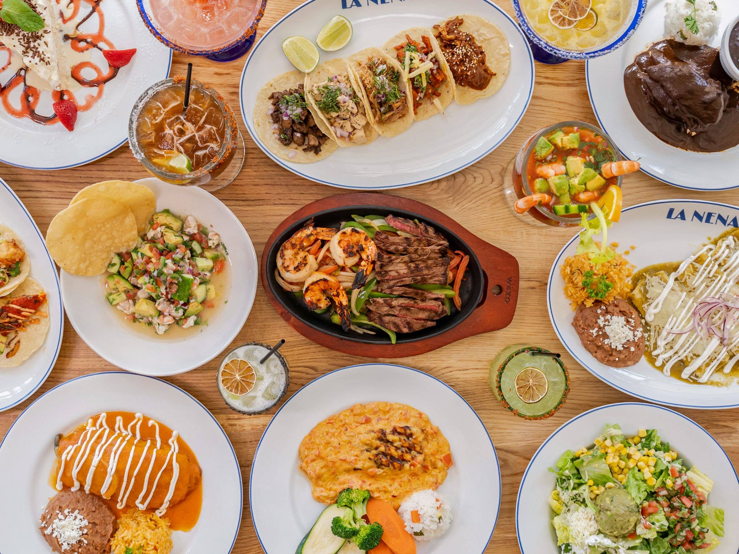Assorted Mexican dishes on a wooden table: tacos, grilled steak, enchiladas, chips, beans, and drinks with lime.