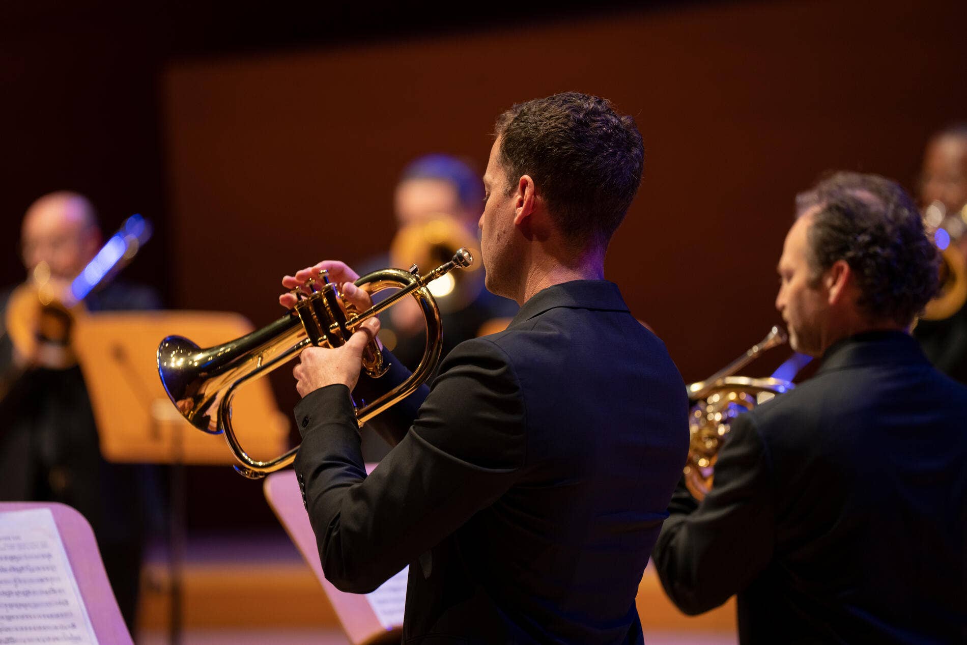 Musician in a concert setting playing a brass trumpet with band members in soft-focus background.