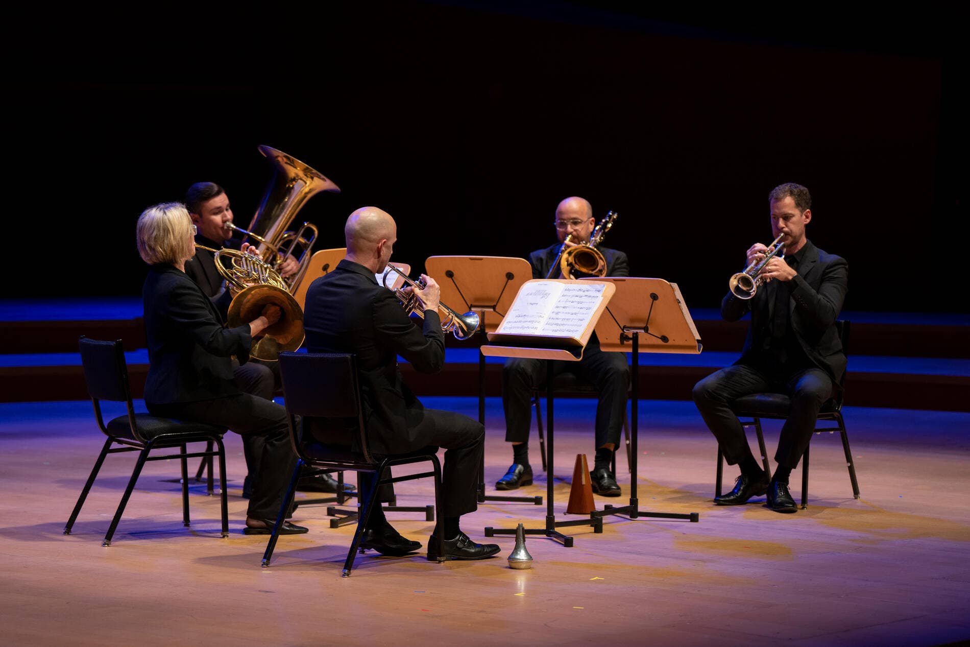 Orchestra brass section seated on stage, playing French horns and trumpet under blue spotlights.