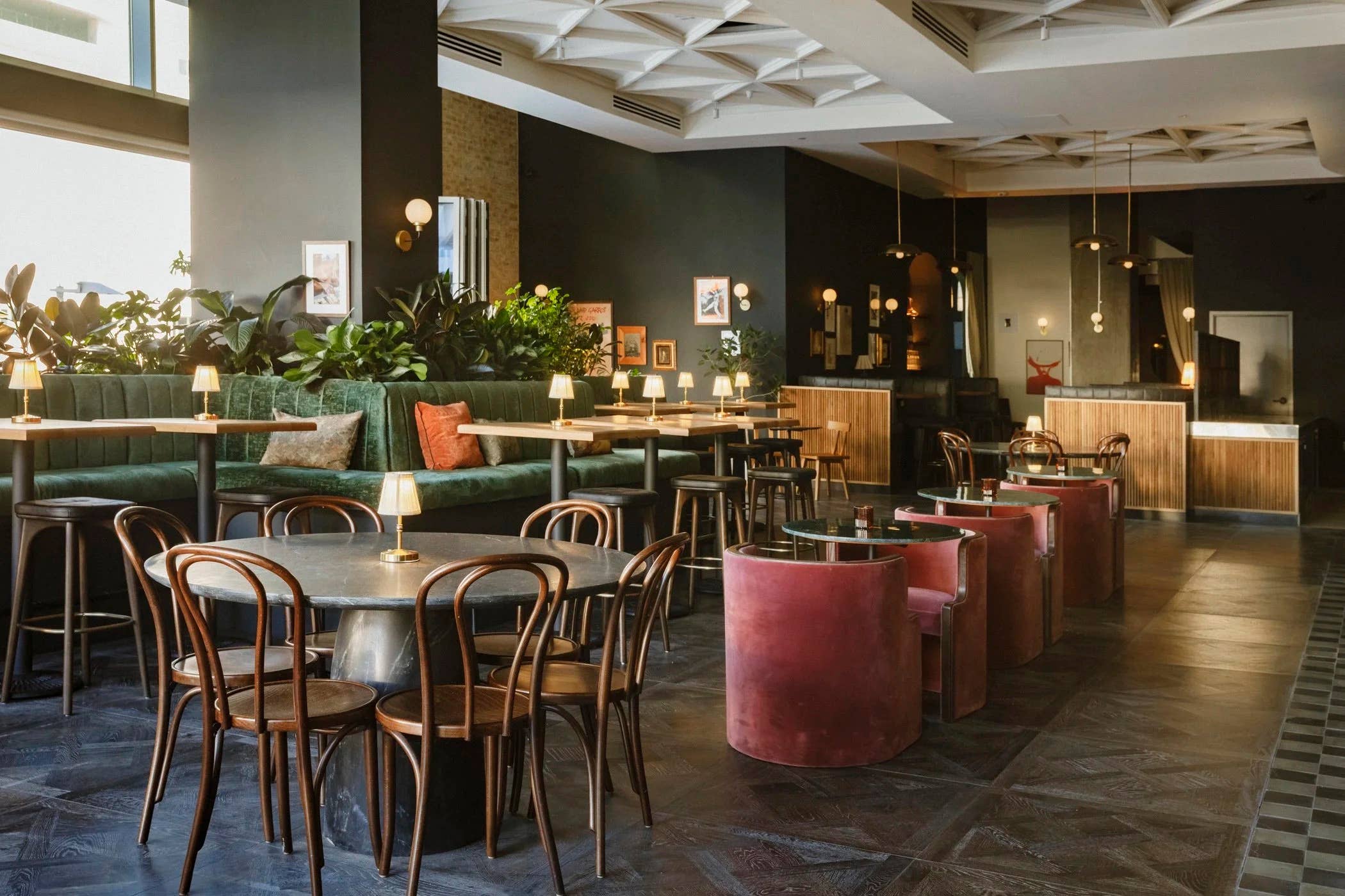 Warm modern lounge with green banquettes, copper chairs, hanging lights, and potted plants under white beams