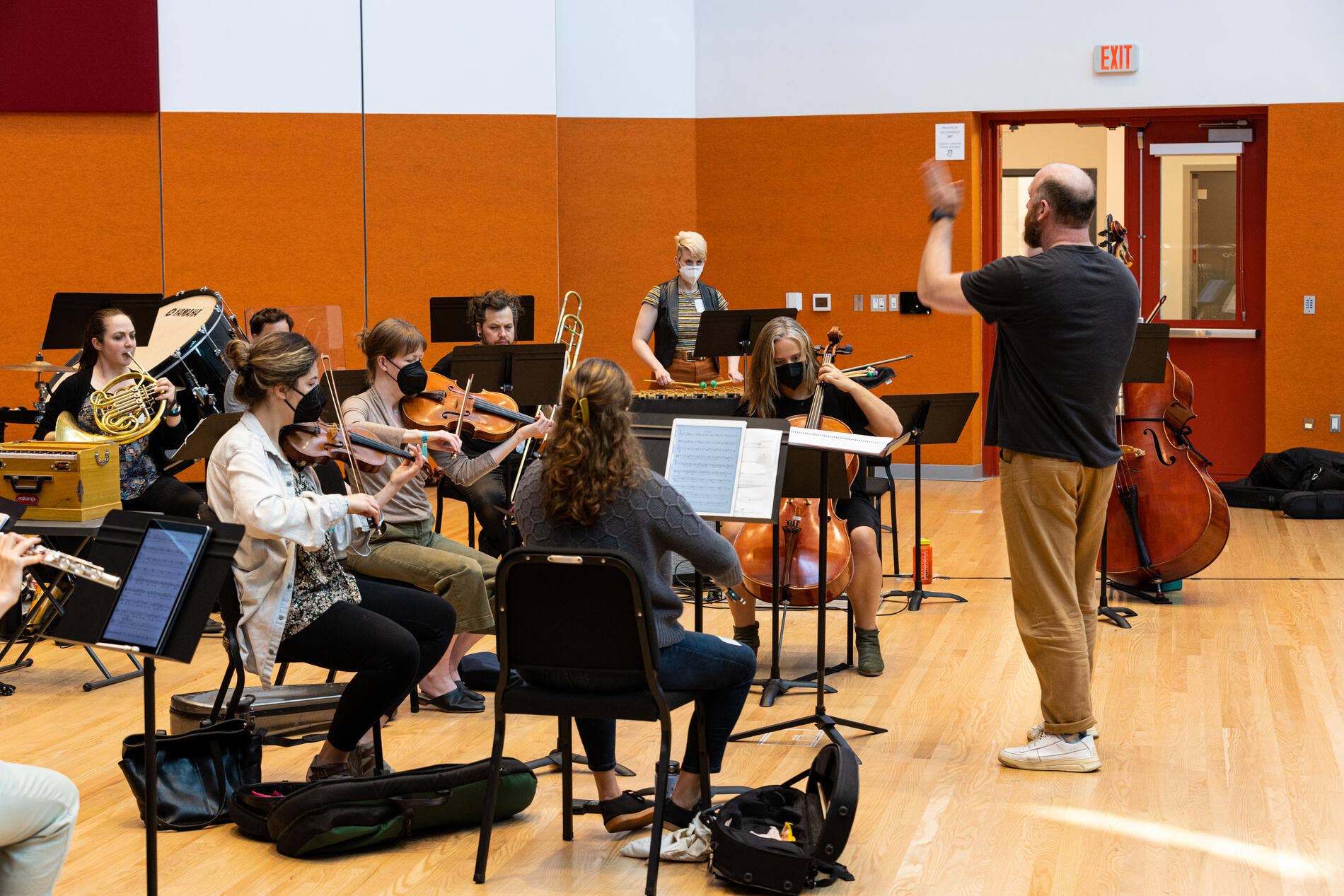 Orchestra rehearsal in a music room with musicians playing strings and a conductor in front, some wearing masks