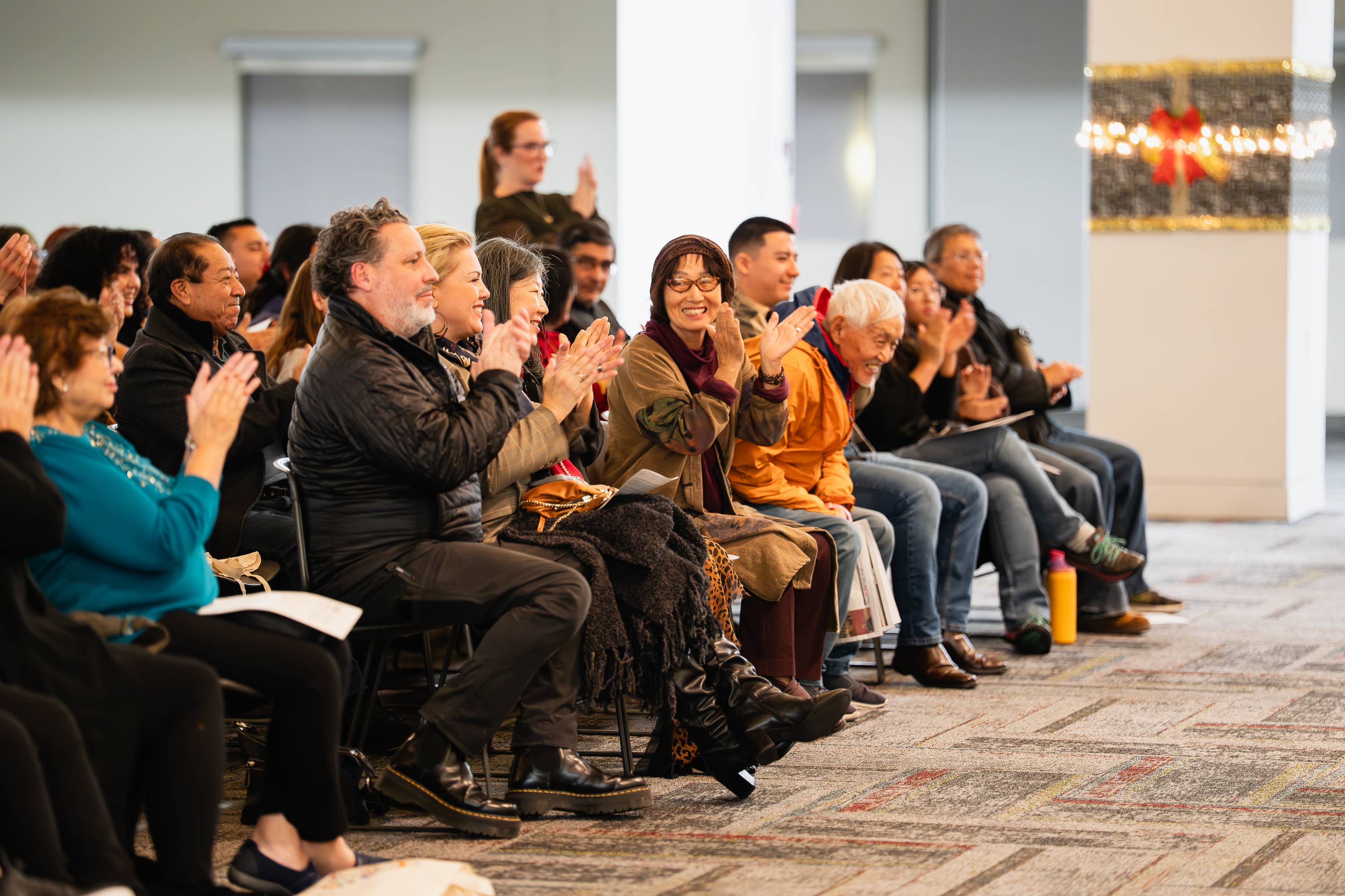 Audience members applauding in an indoor hall, seated rows facing the stage, with a speaker in the background.