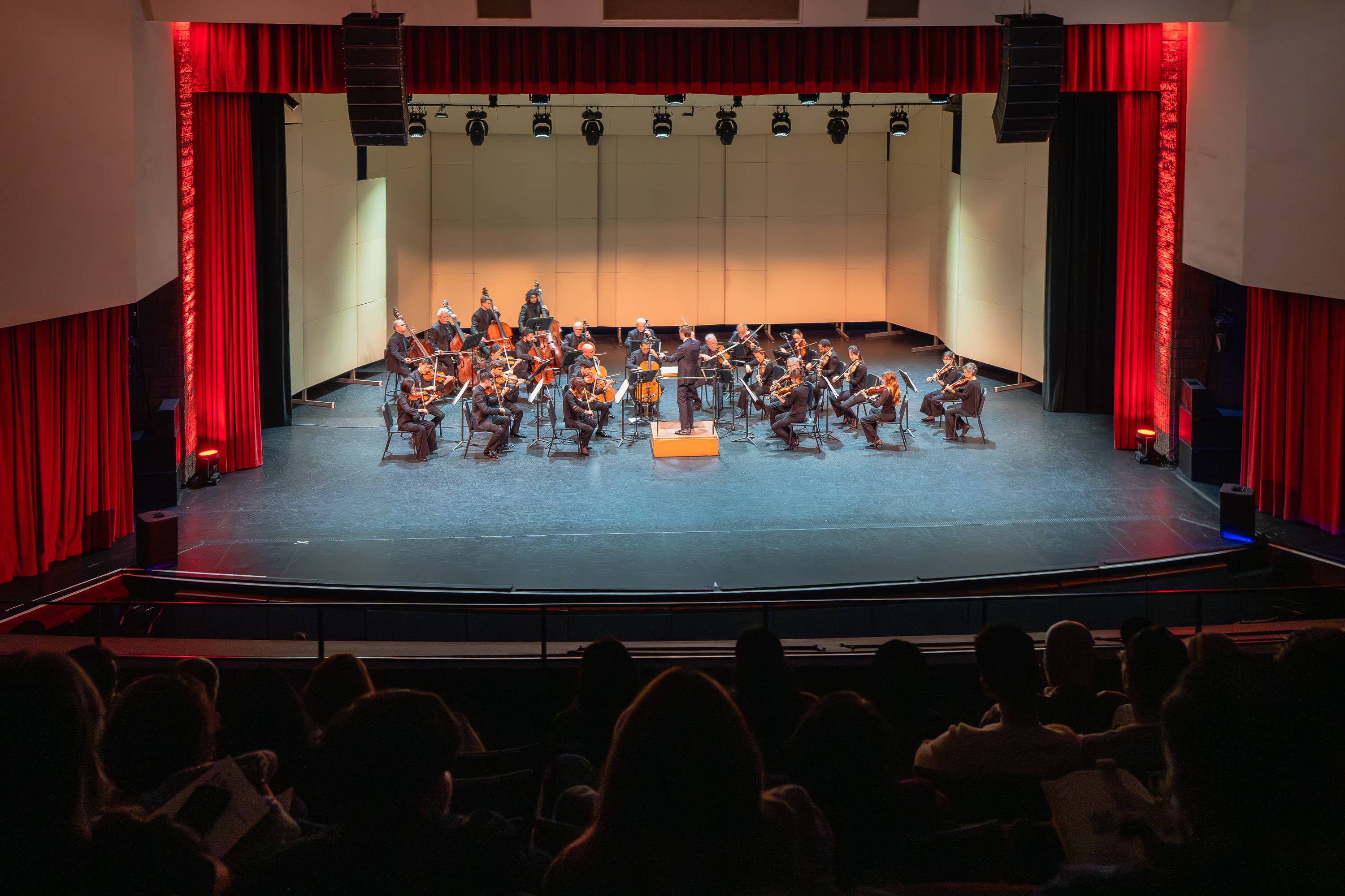 Orchestra seated on a stage under red curtains, strings and brass performing before an empty backdrop.