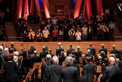 Orchestra audience applauds as performers stand center stage under red and blue spotlights in a concert hall