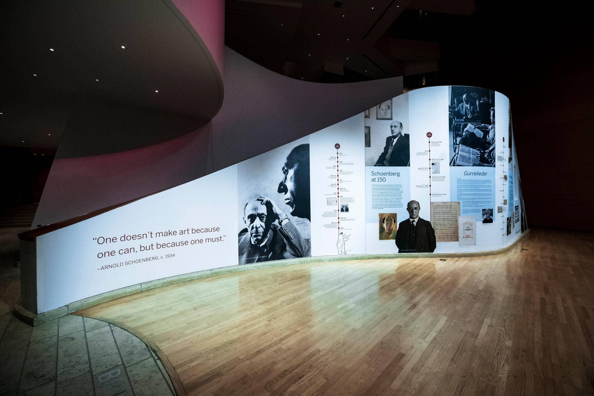 Curved museum exhibit wall with portraits and text about Schoenberg and his music, in a dark gallery.