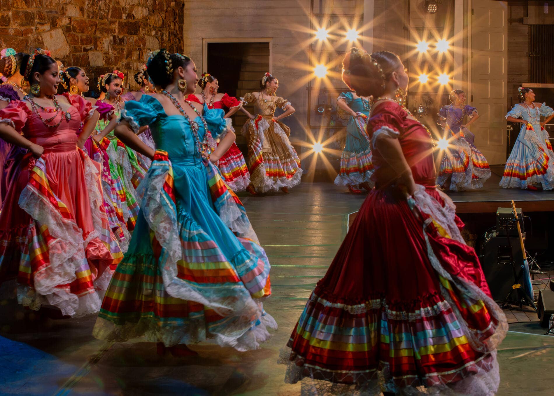 Dancers in colorful folkloric dresses perform on a stage, lit by bright spotlights and glowing beams.