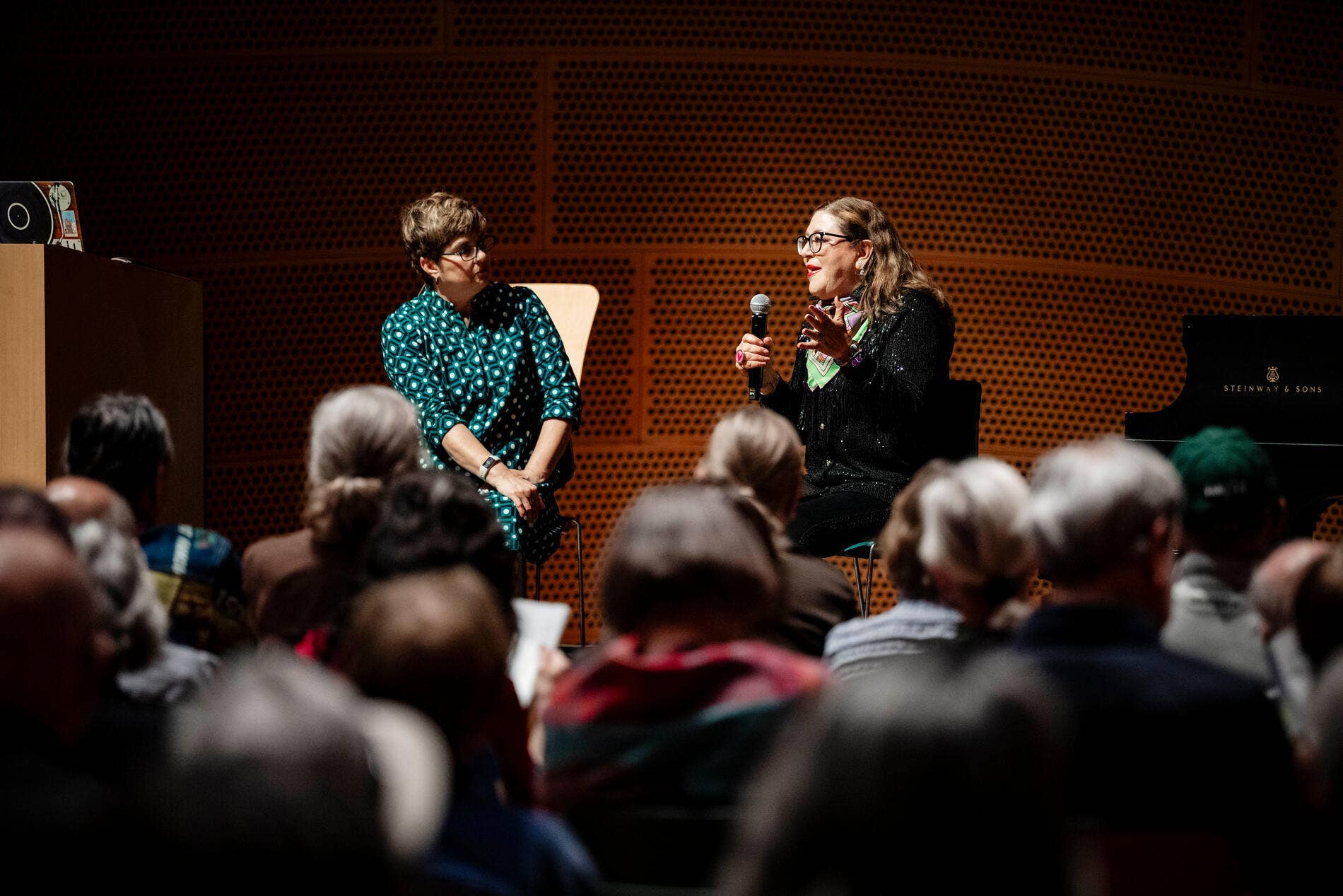 Two women sit on stage, one holding a microphone, addressing an audience at an indoor event.