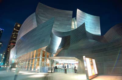 Walt Disney Concert Hall's modern curved museum building with glass entrance lights at night, surrounded by tall city towers