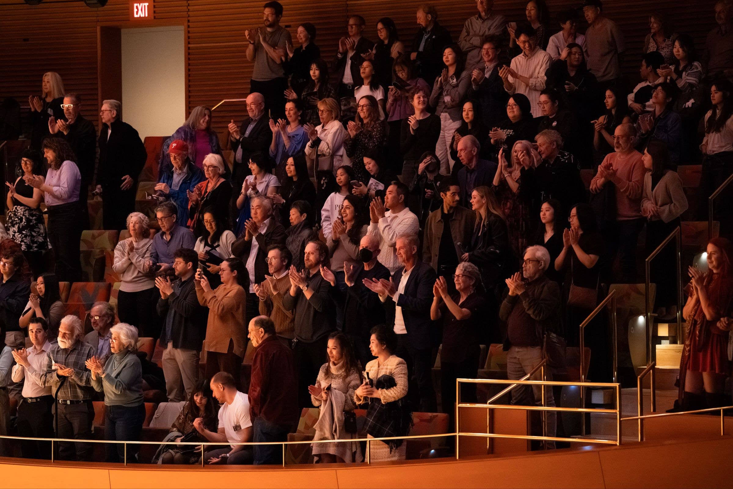 A large audience in a theater clap and cheer under warm red-orange lighting, exit sign glowing in back