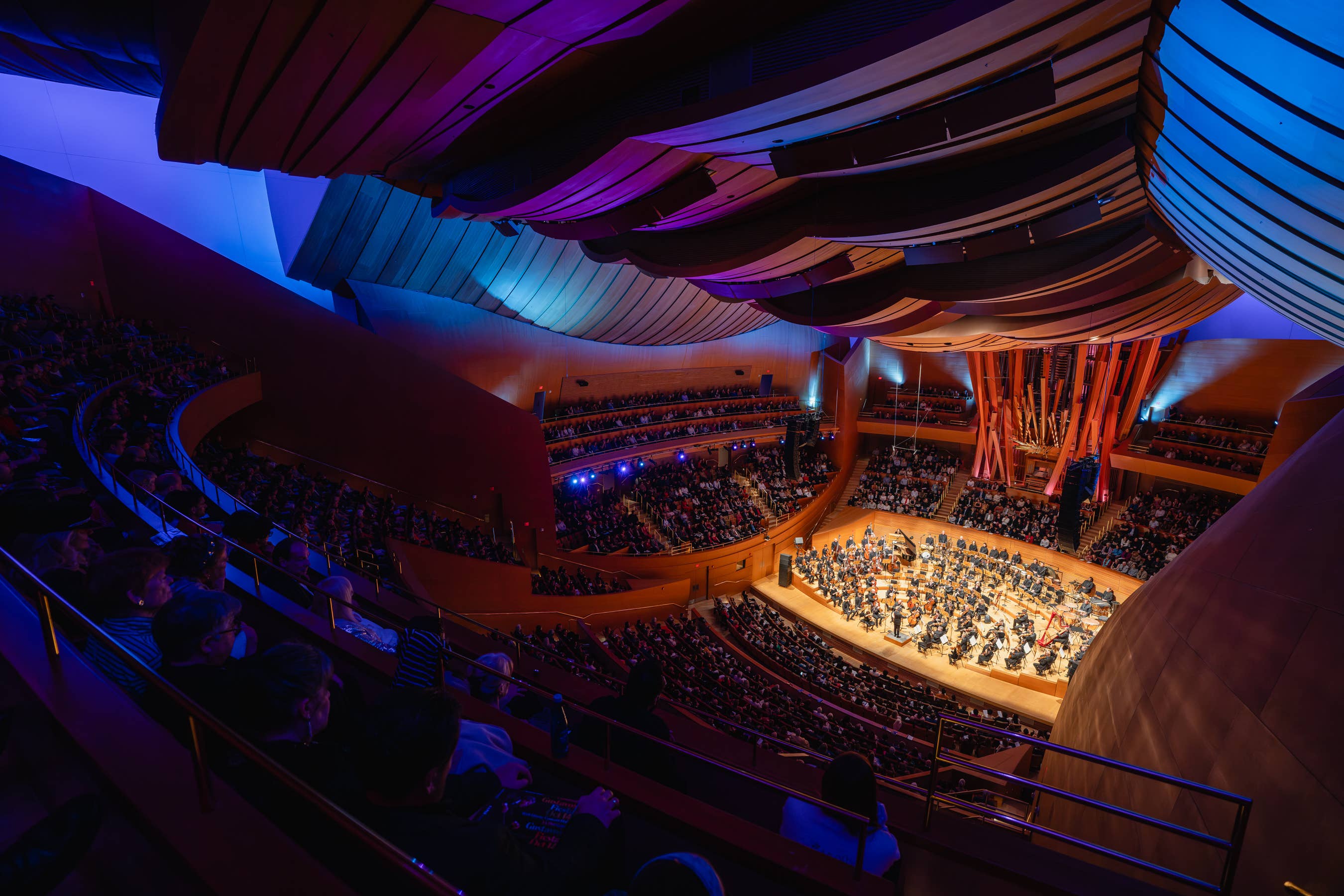 Wide view of an illuminated concert hall with an orchestra on stage and audiences in multi-level balconies.