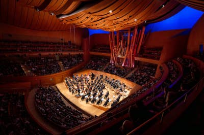 Concert hall interior with an orchestra performing onstage under warm lights, seen from an upper balcony.