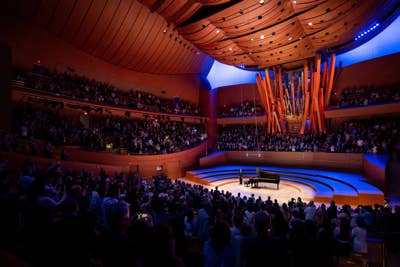 El público llena la sala de conciertos mientras un ponente se encuentra en el escenario, frente a un piano de cola, piano una iluminación azul.