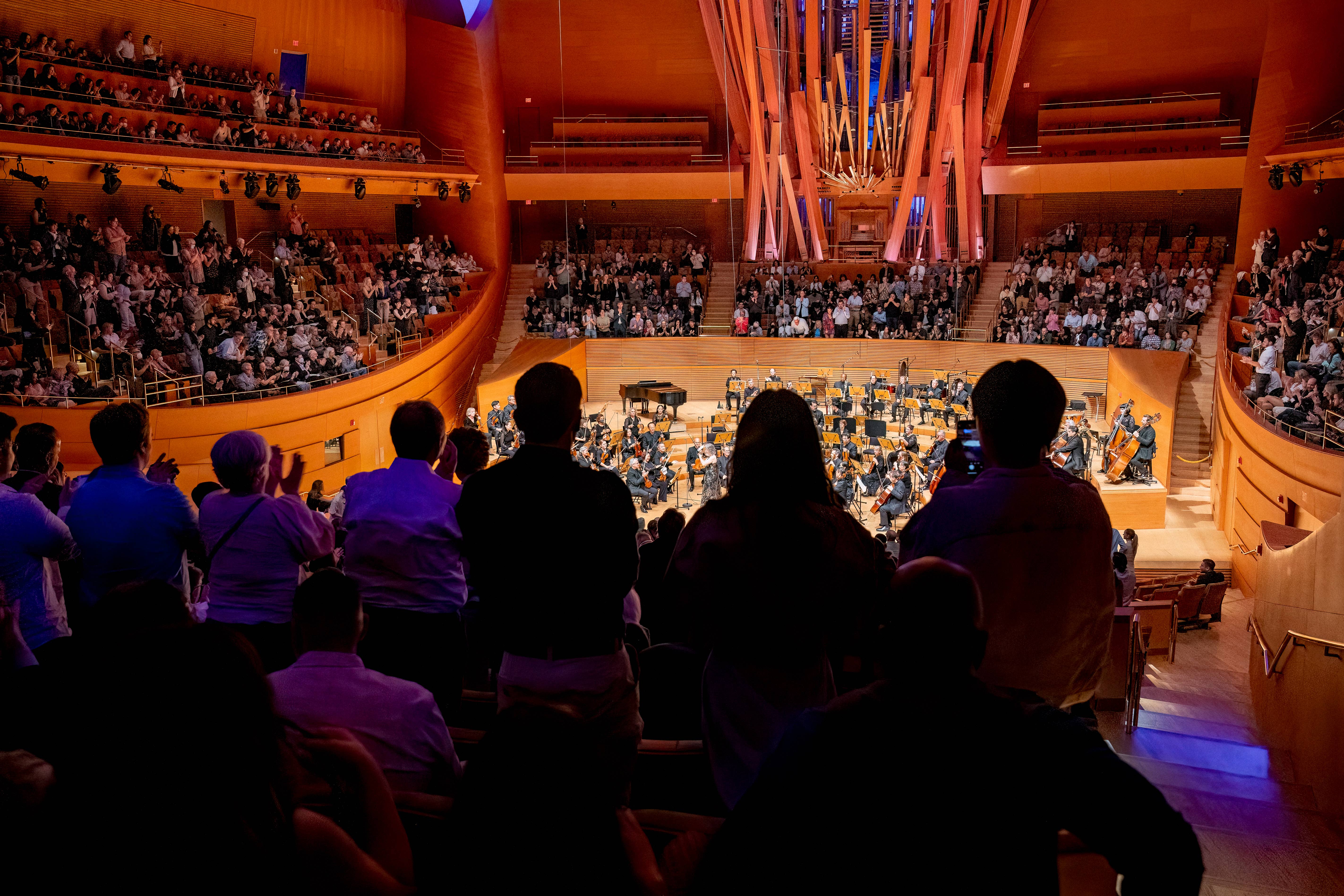 Audience applauds as a large orchestra performs on a lit concert stage inside a grand auditorium.