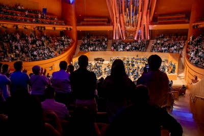 Audience applauds as a large orchestra performs on a lit concert stage inside a grand auditorium.