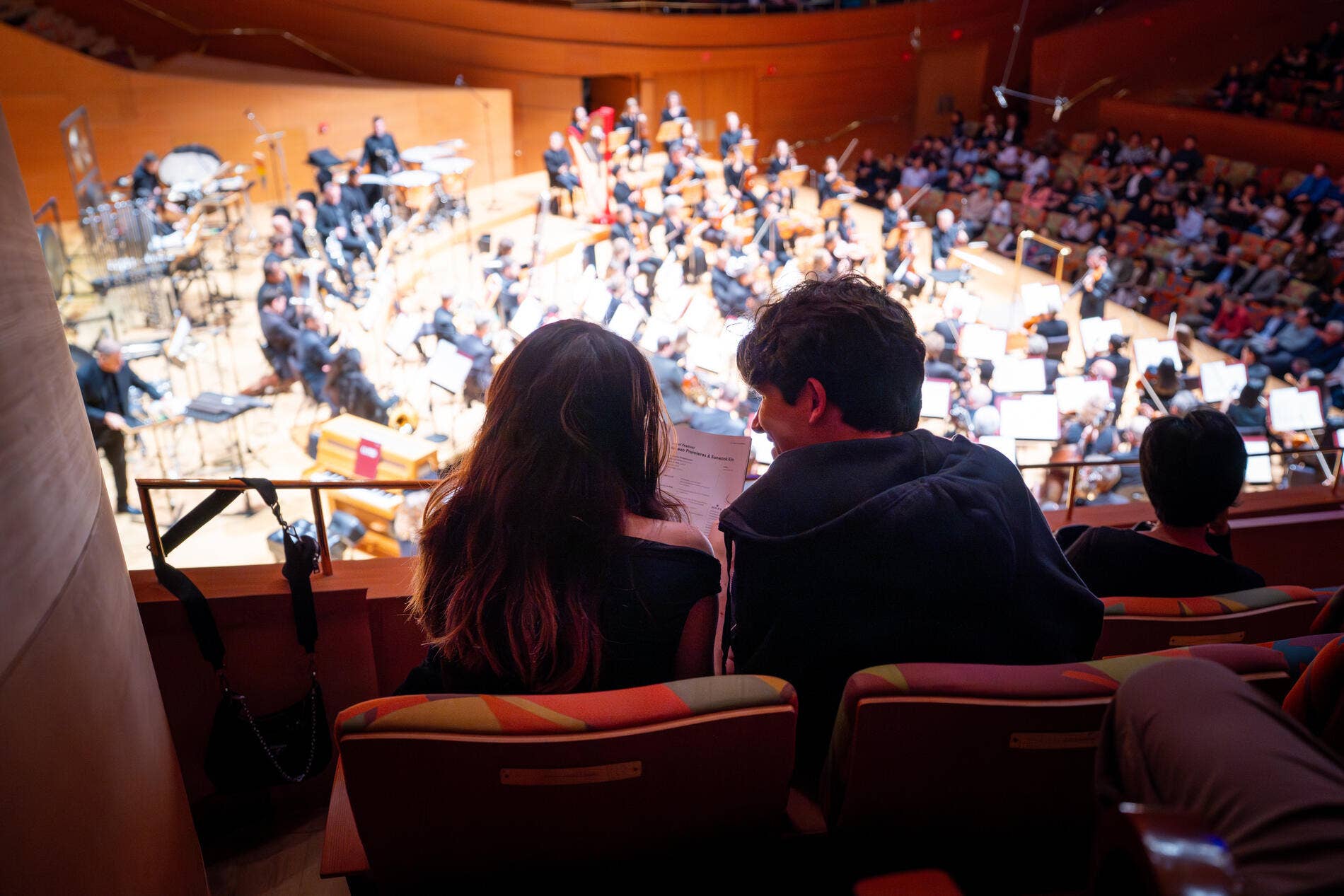 Audience sits in theater seats watching an orchestra with instruments on stage under warm lights.