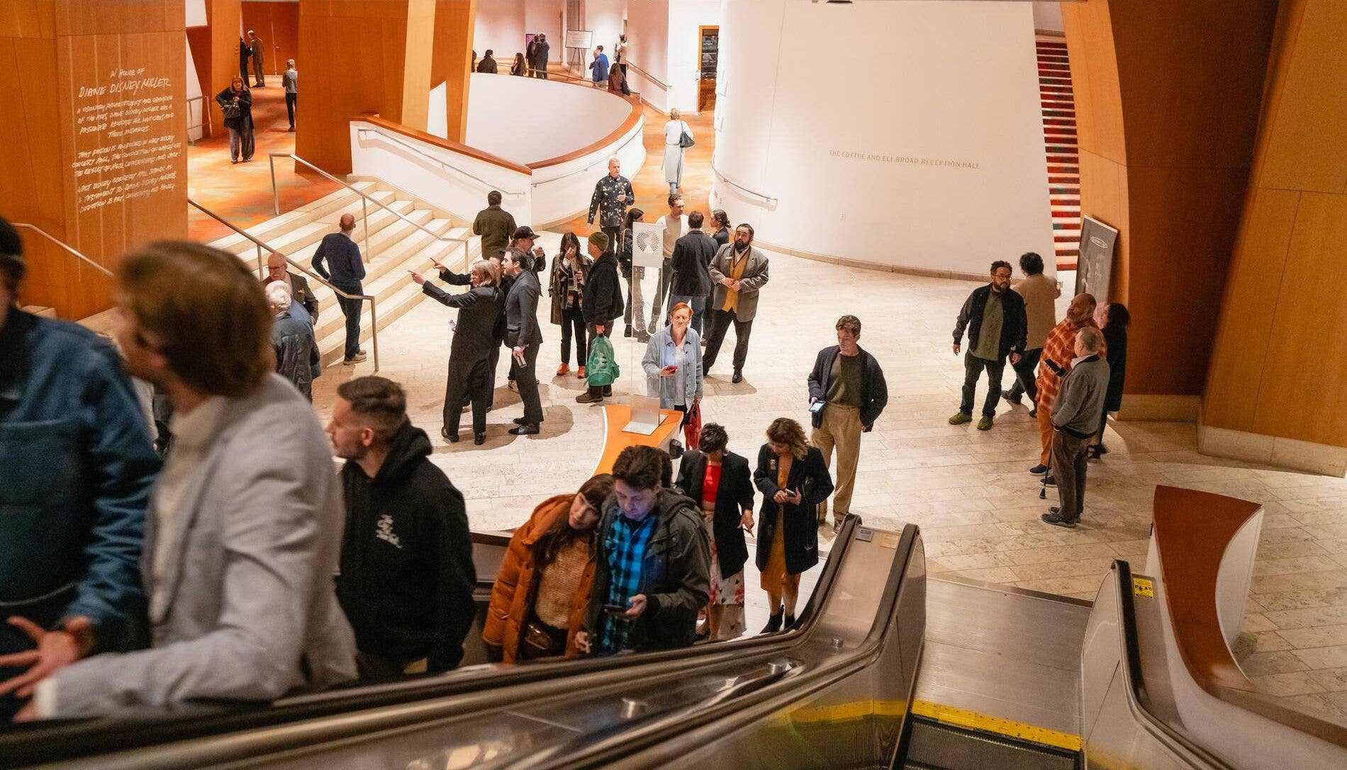 People mingle in a modern museum lobby with a curved staircase, art displays, and warm interior lighting.