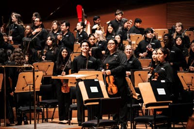 YOLA orchestra members in black uniforms perform onstage with instruments and red flags behind them.