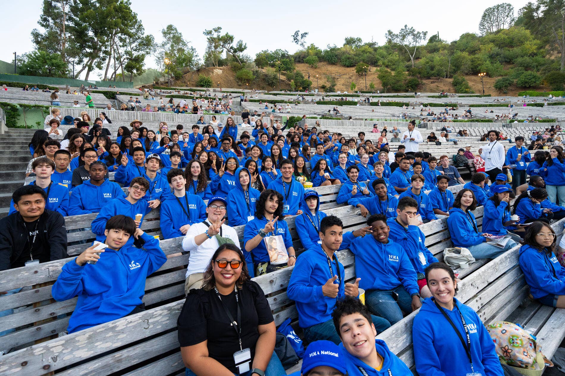 Large group of students in blue hoodies posing on outdoor amphitheater benches, smiling and making peace signs.