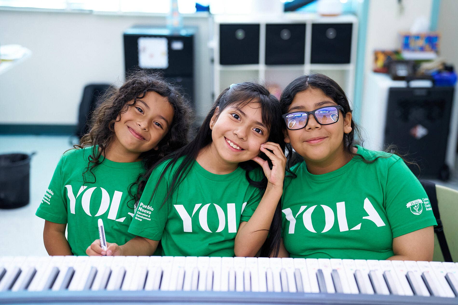 Three girls in green YOLA shirts posing together, one wearing blue reflective glasses, in a classroom.