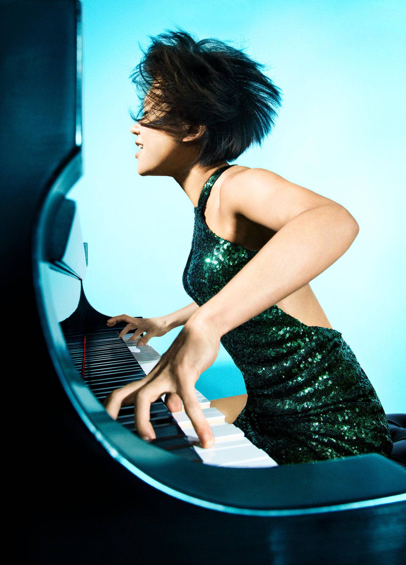 Yuja Wang in a green sequin dress plays a black-and-white keyboard under blue lighting, viewed in profile.