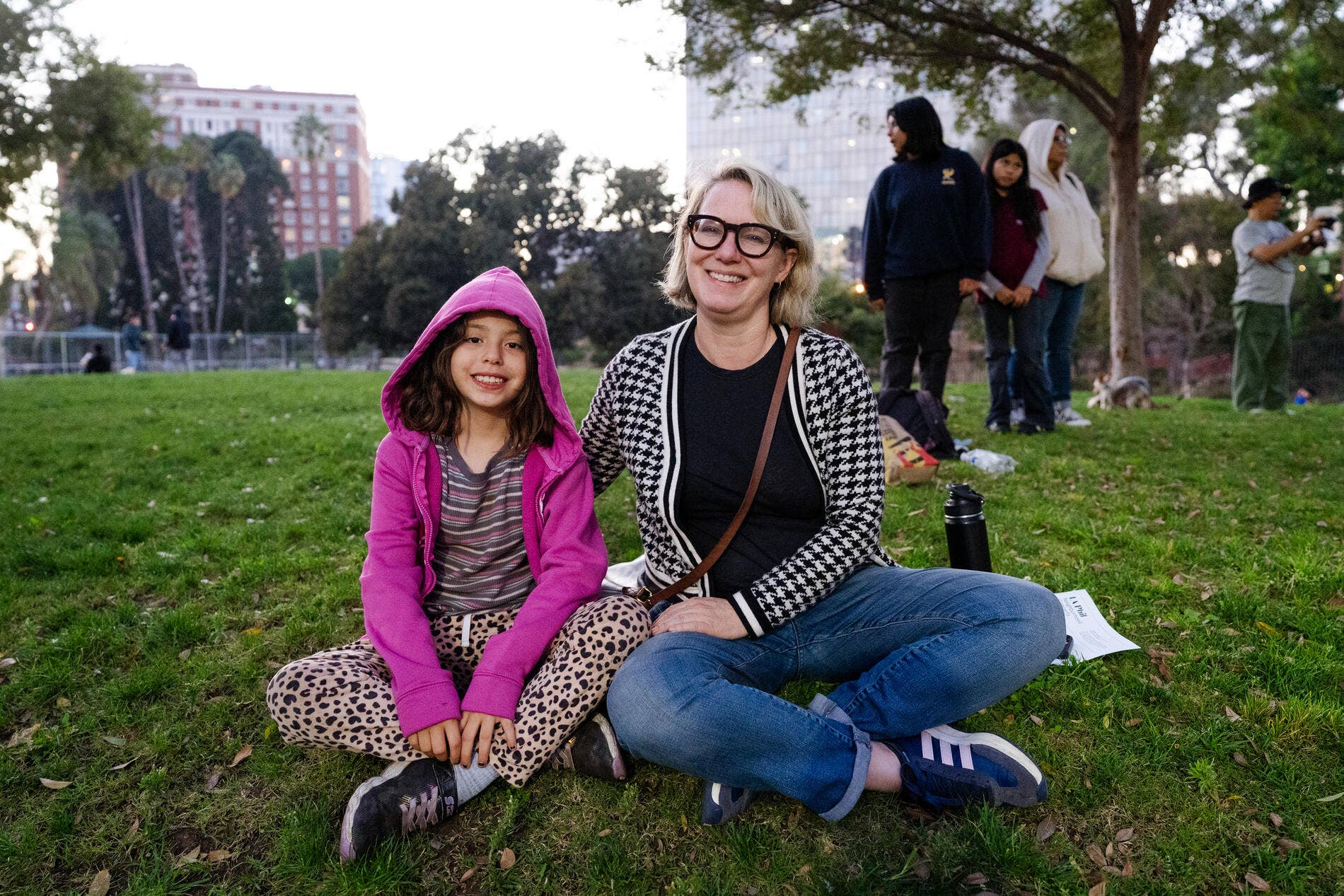 Smiling woman and child in park on grass, with buildings and other people in the background.