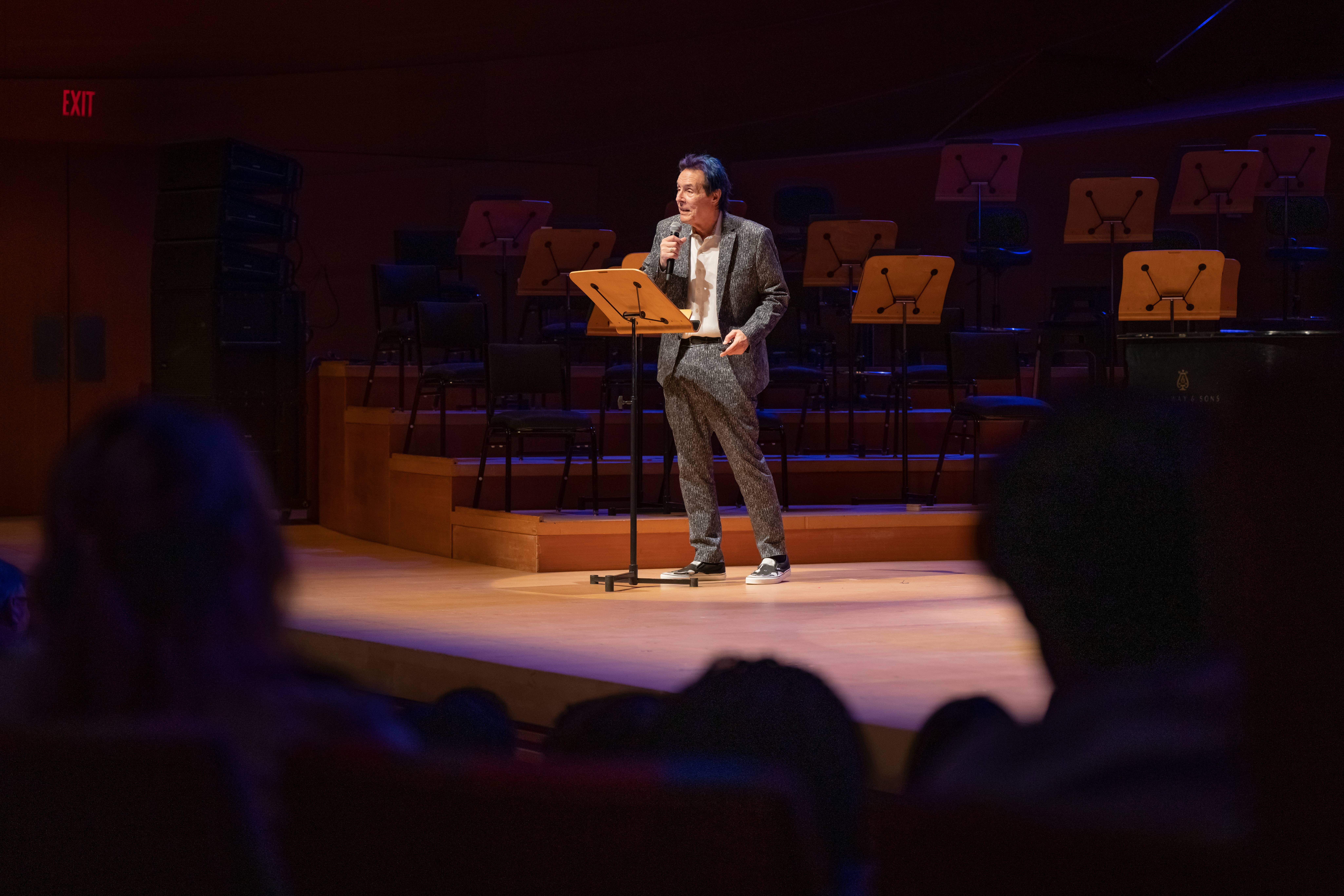 Man in a suit speaks into a microphone on stage beside a music stand during a live event.