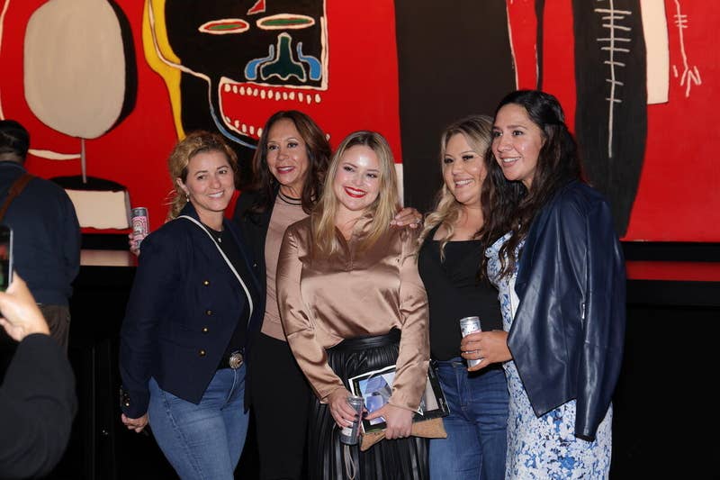 Group of women posing with awards and drinks at an event in front of a colorful mural background