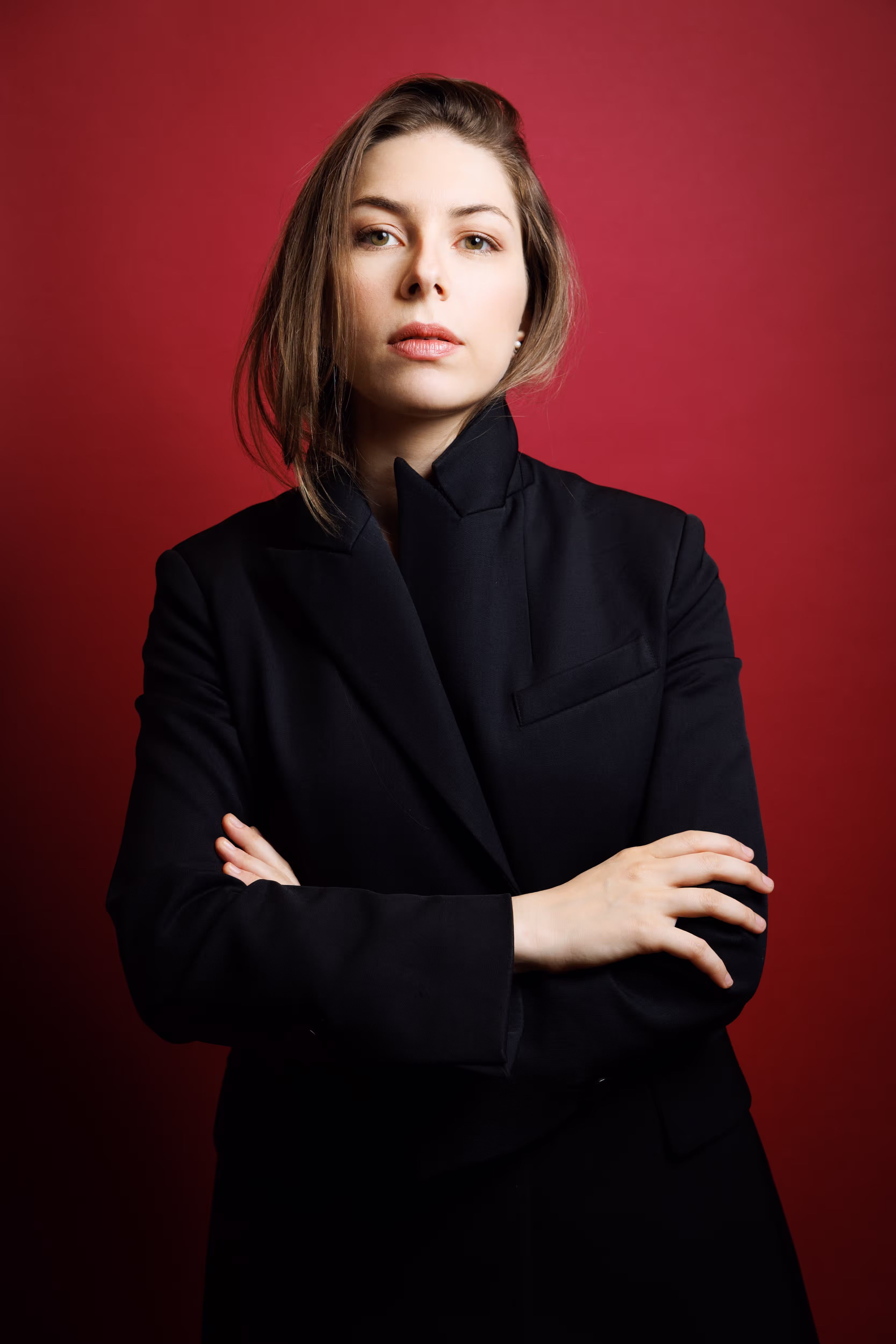Professional headshot of a woman with arms crossed, wearing a black blazer against a red background.
