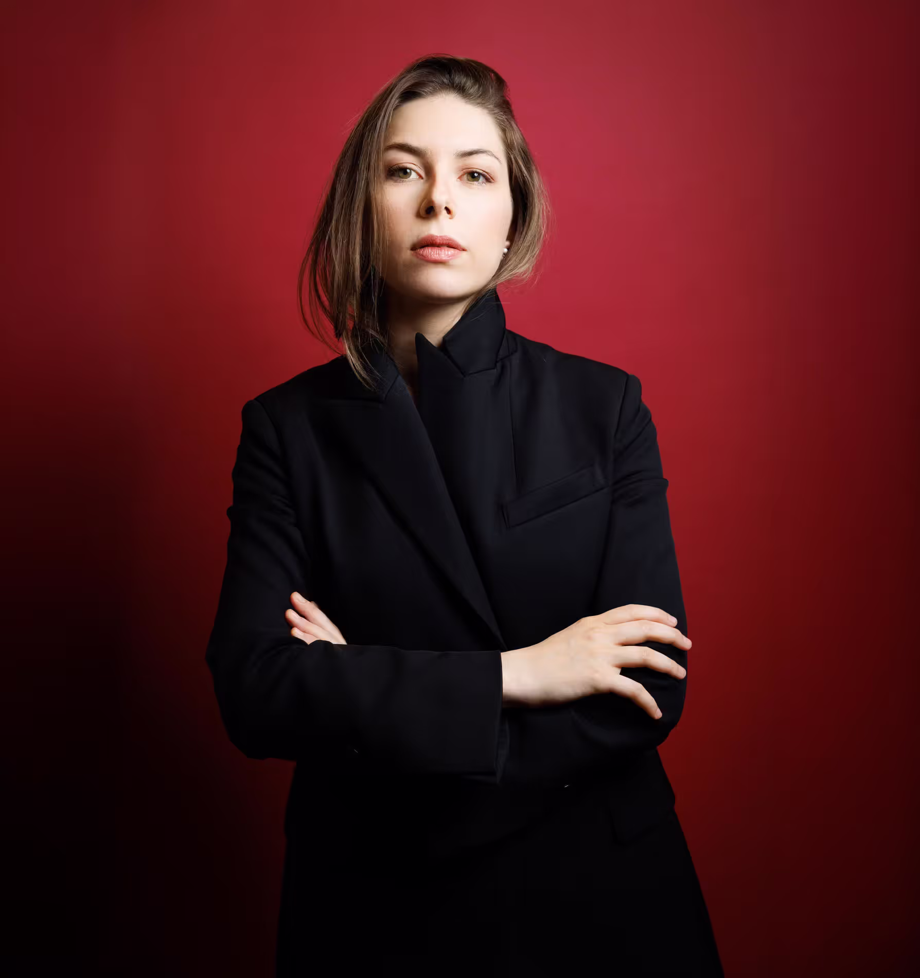 Anna Handler in a black blazer with arms crossed, looking at camera against a red studio background.