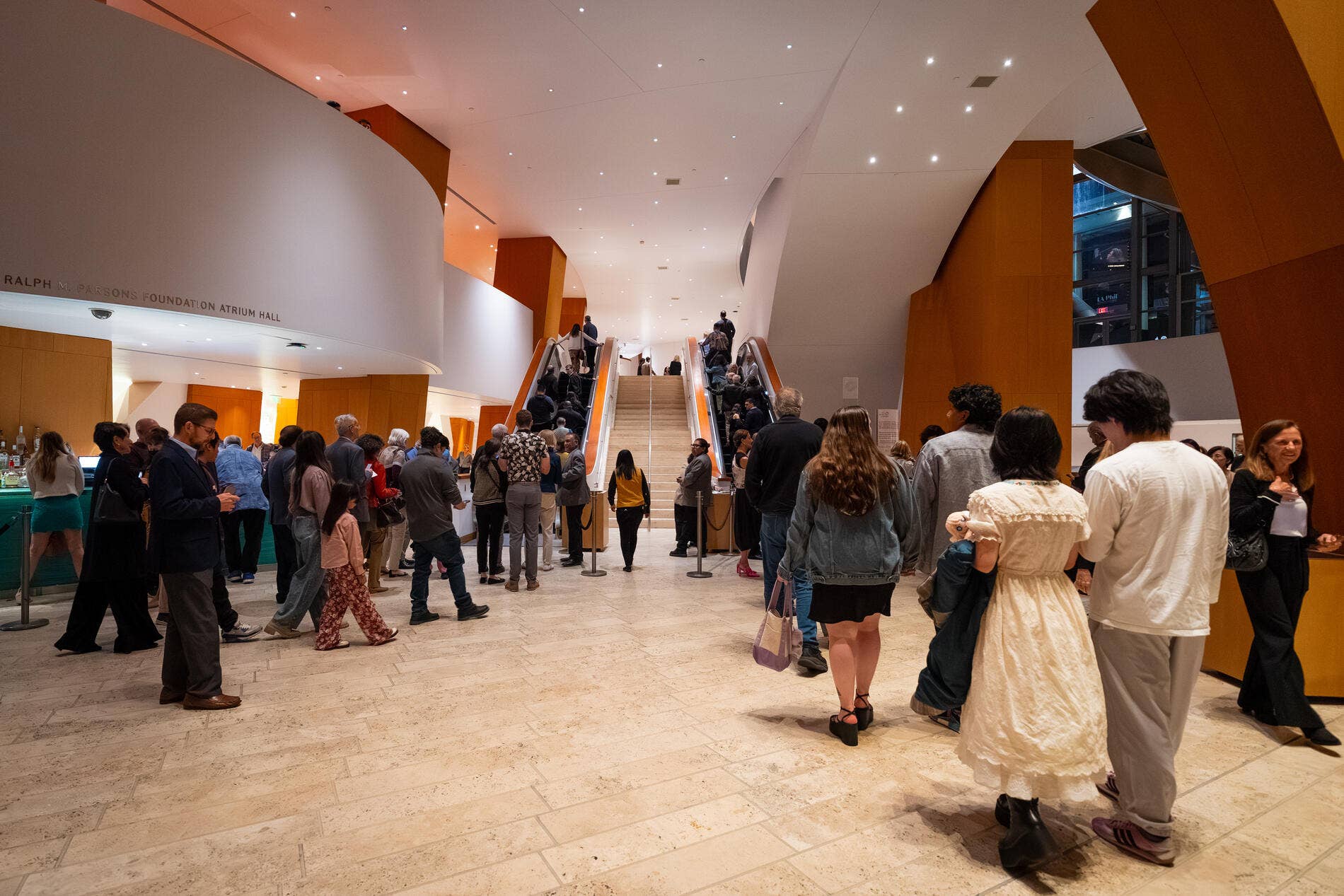 Crowd gathers in a modern museum atrium with tall curved walls, glowing ceiling lights, and escalator stairs.