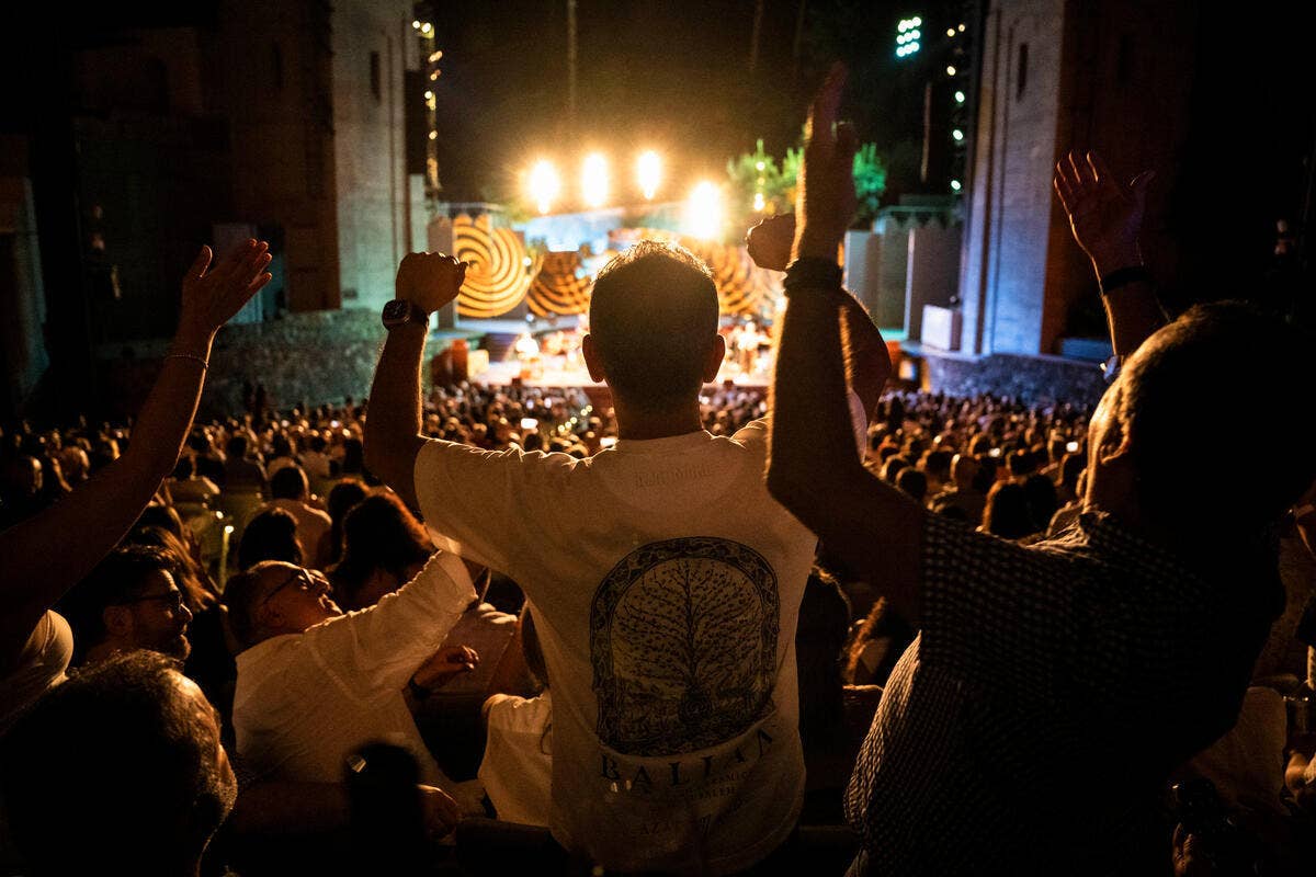 Crowd cheers at night concert, silhouetted fans raising arms toward bright stage lights and speakers.