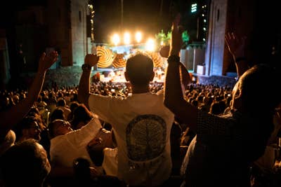 Crowd cheers at night concert, silhouetted fans raising arms toward bright stage lights and speakers.