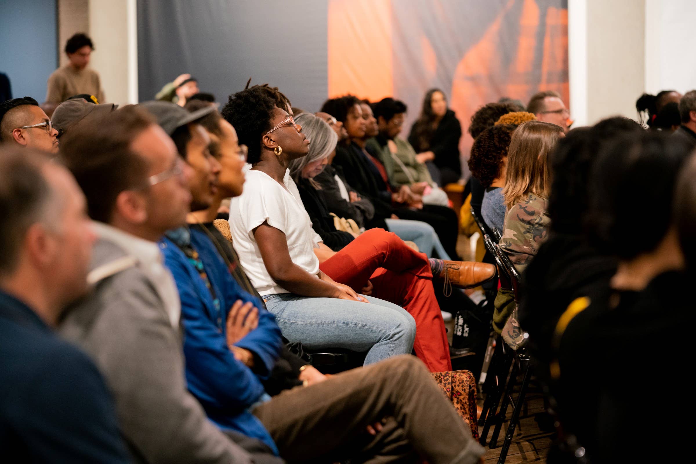 Audience members seated in a conference room, listening attentively with microphones and stage lights in back.