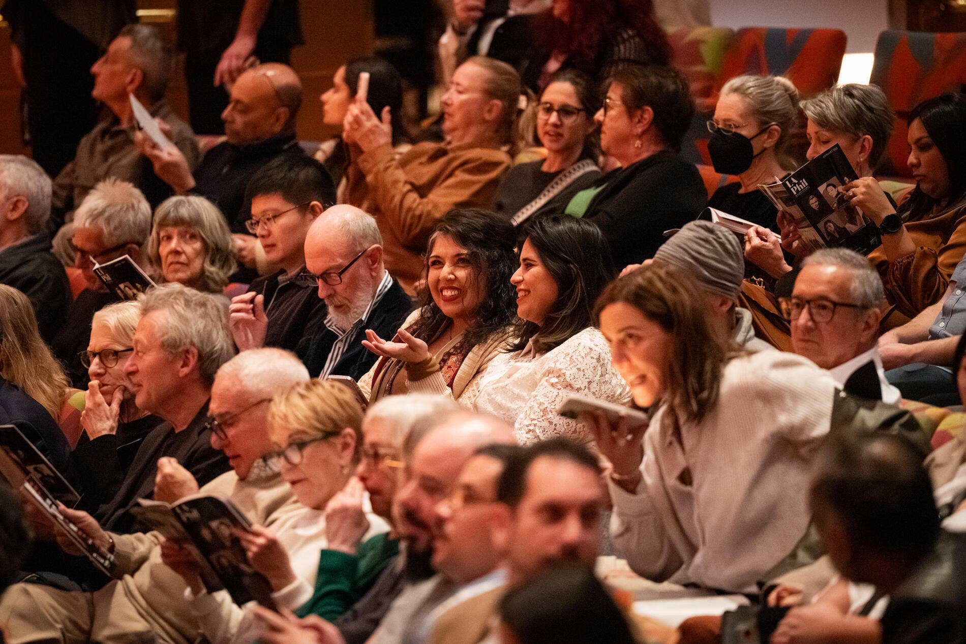 Audience applauds in an indoor theater, with people seated and some wearing black face masks and holding programs.