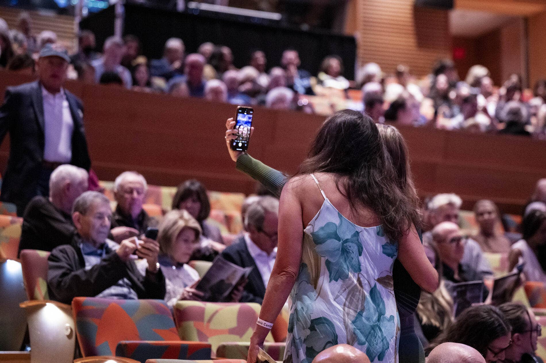 Woman in floral dress raises smartphone to film a seated audience at a conference or event.