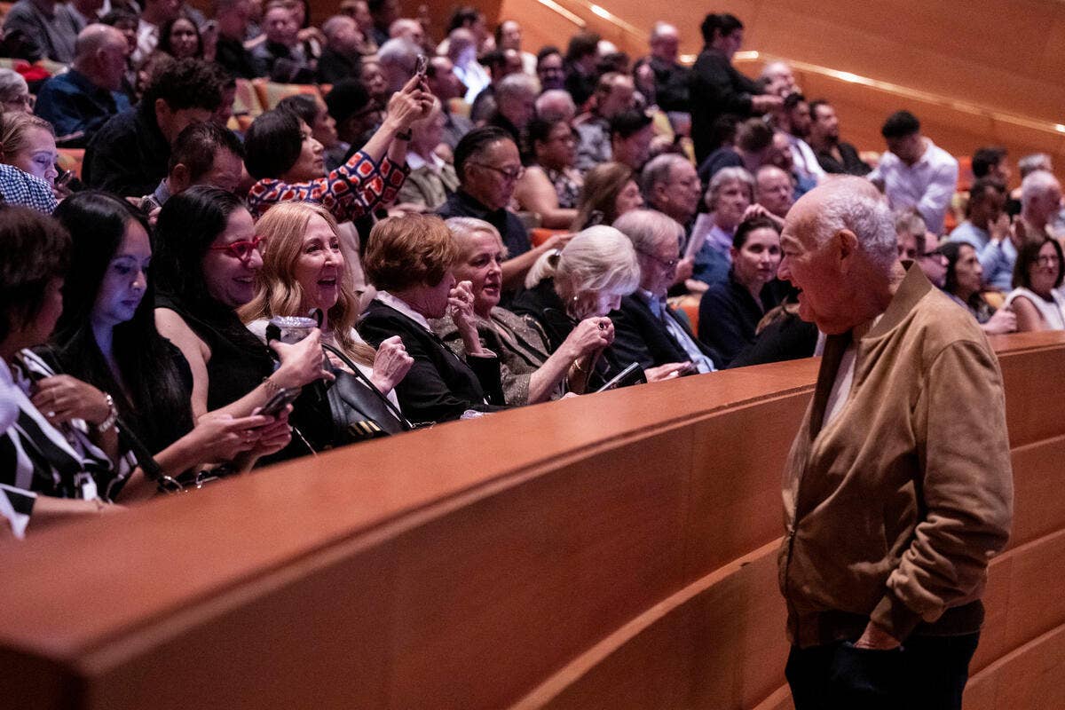Elderly man speaks to an applauding crowd in a conference hall, with audience members clapping and smiling