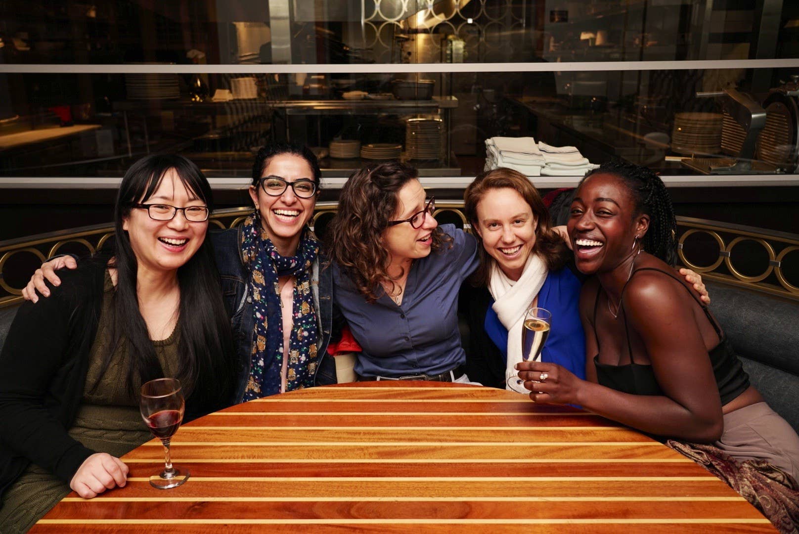 Five friends sit at a restaurant table, laughing together with glasses of champagne.