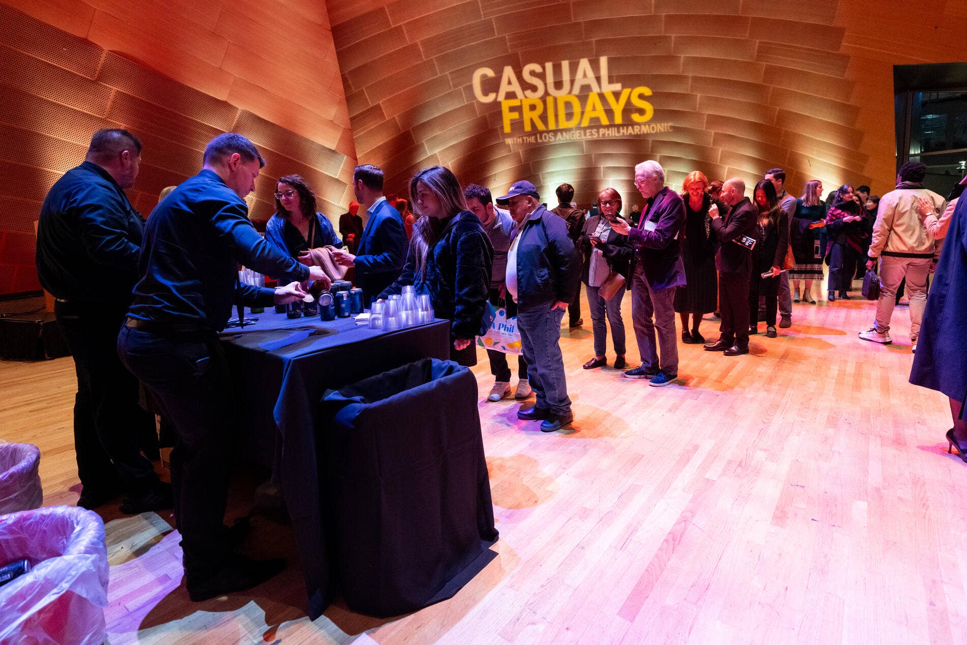 Volunteers serve drinks at a “Casual Fridays” event with Los Angeles Philharmonic backdrop in a concert hall.