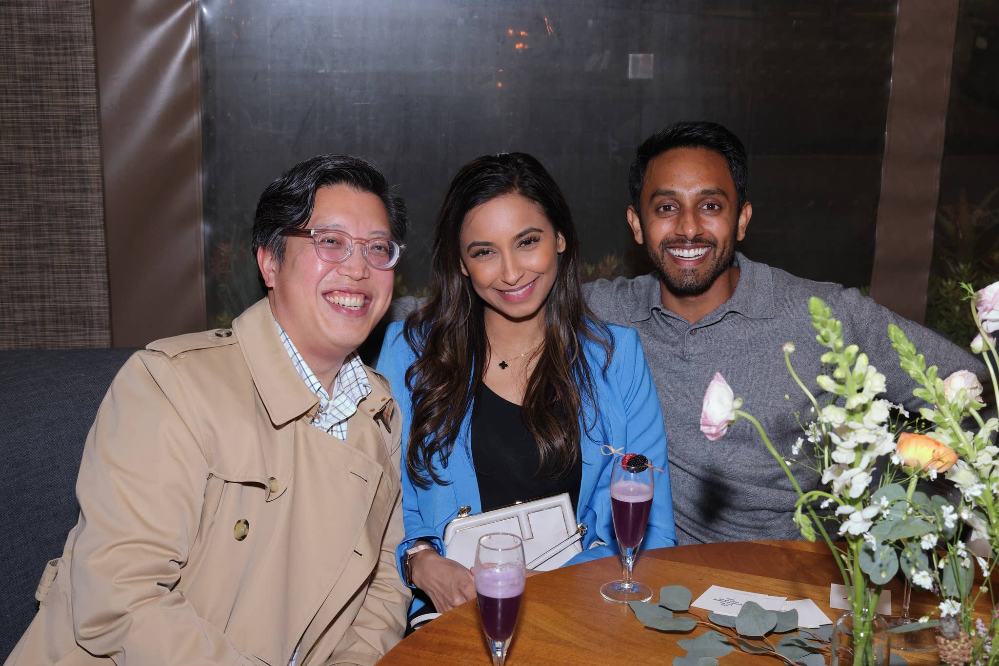 Smiling trio seated at a banquet table with drinks and floral centerpiece against a dark wall backdrop