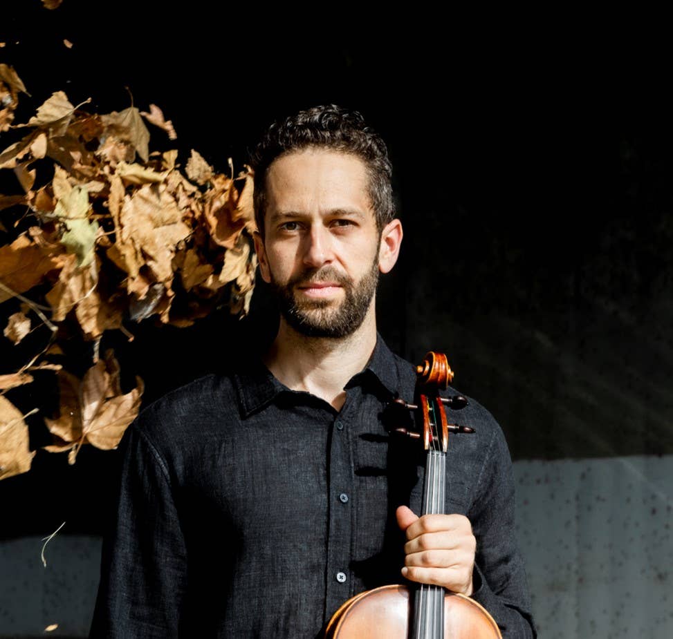 Man holding a violin, wearing a dark shirt, with dry leaves on the left against a dark background.