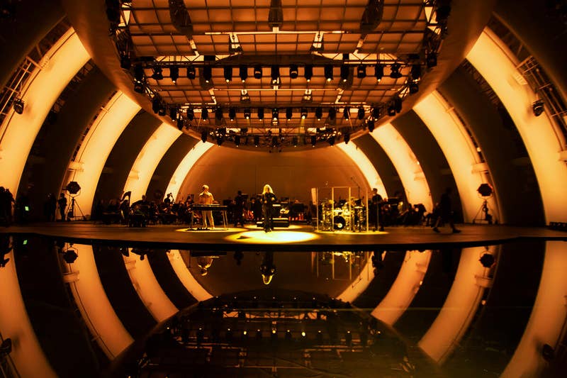Concert stage with musicians and glowing lights overhead, with drum kit and performers under warm yellow illumination.