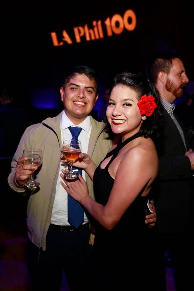 Two people smiling at a party, holding champagne flutes, woman wearing a red flower in her hair.